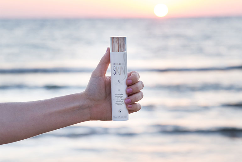Kvinde ved havet, der står med et Herbalife SKIN-sommerhudplejeprodukt